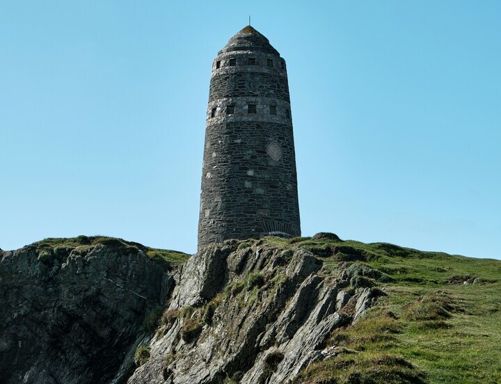Historic cylindrical stone tower with narrow slit windows perched on a rugged coastal cliff, green grass under clear blue sky