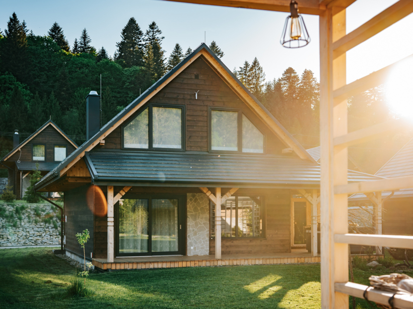 Drewniany domek wypoczynkowy z dużymi oknami i tarasem, otoczony zielonym trawnikiem i lasem, przy zachodzie słońca