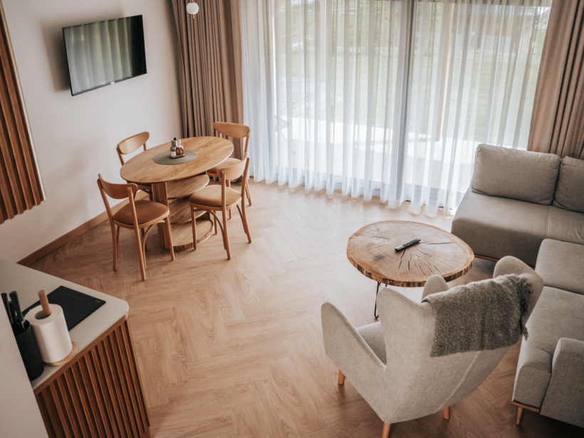 Przestronny pokój dzienny z szarą sofą, fotelem, drewnianym stołem i krzesłami, oraz dużymi zasłonami przepuszczającymi naturalne światło