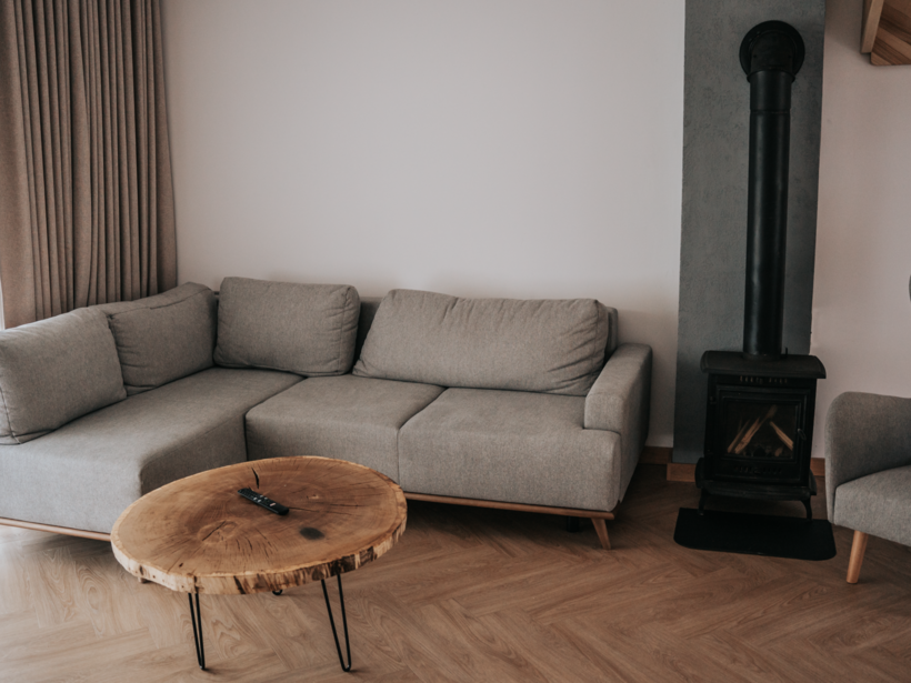 Przytulny salon z szarą sofą narożną, stołem z naturalnego drewna i kominkiem na drewno, jasne zasłony i drewniana podłoga.