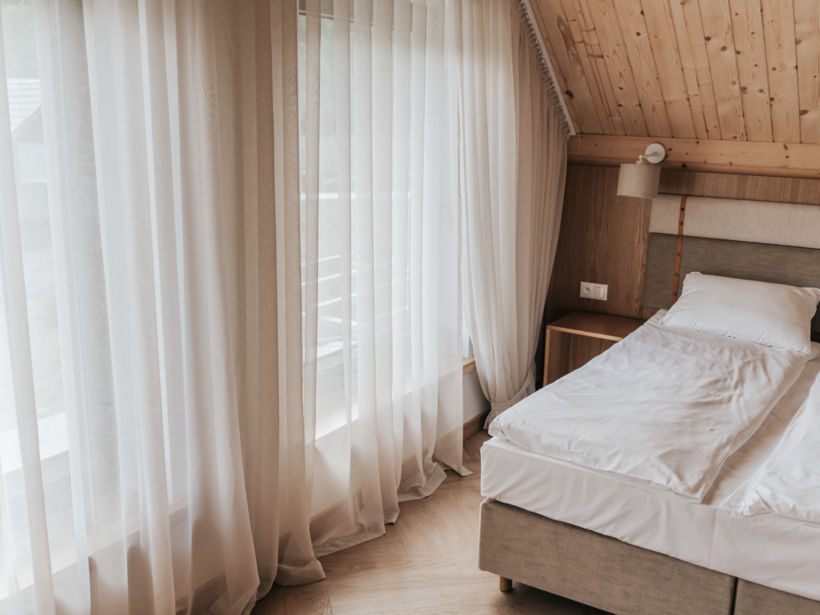 Pokój hotelowy z drewnianym sufitem, dużym łóżkiem z białą pościelą i zasłonami wpuszczającymi naturalne światło
