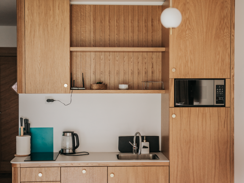 Kuchnia w pokoju hotelowym z drewnianymi szafkami, zlewem, czajnikiem elektrycznym i mikrofalówką, przytulna jadalnia z drewnianym stołem