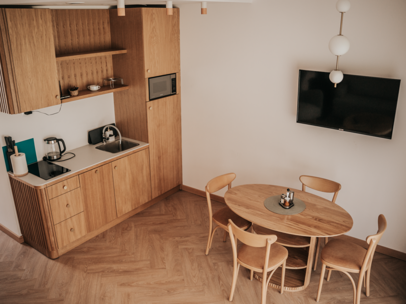 Kompaktowy aneks kuchenny z drewnianymi szafkami, mikrofalówką, czajnikiem, stołem i czterema krzesłami w przytulnym wnętrzu apartamentu