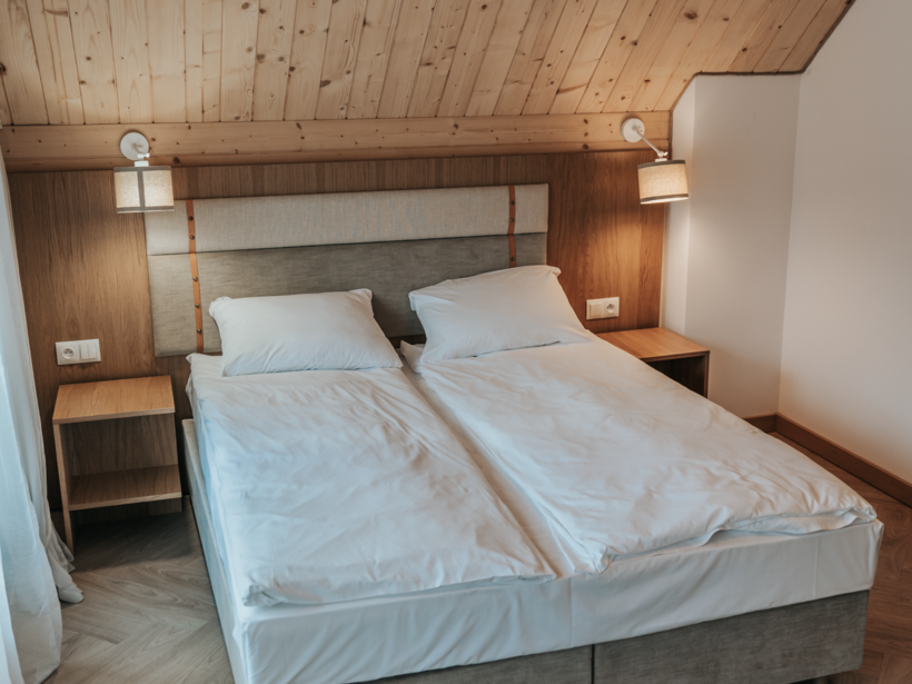 Pokój hotelowy z podwójnym łóżkiem, drewnianym podwieszanym sufitem, lampkami przy łóżku i prostymi stolikami nocnymi