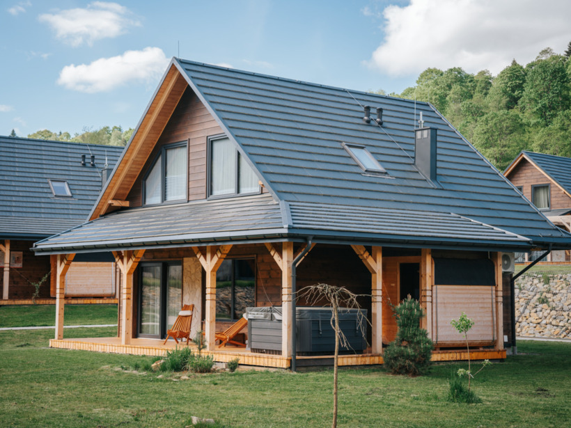 Drewniany domek z tarasem, leżakami i jacuzzi na trawniku, otoczony zielonymi wzgórzami i lasem