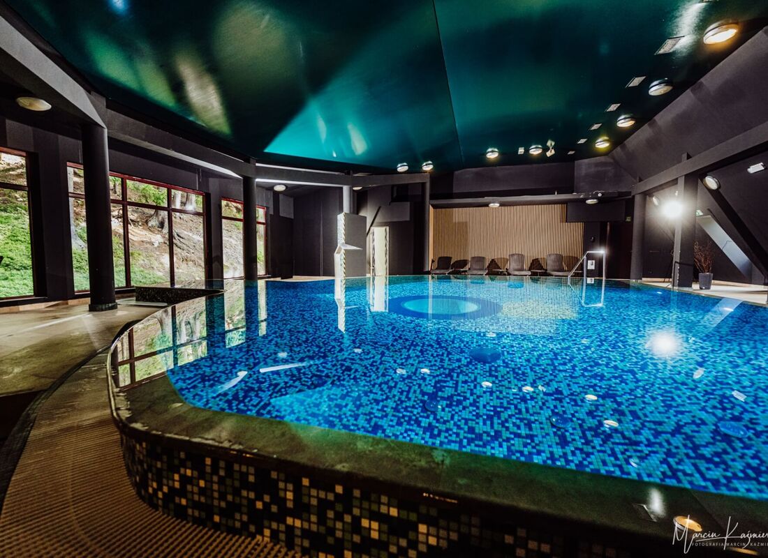 Überdachter Hotelpool mit blauen Mosaikfliesen, geschwungener Kante, Edelstahl-Handlauf, Liegen und großen Fenstern zum bewaldeten Hang