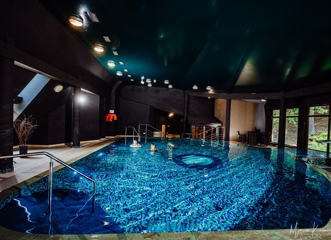 Gemütlicher Indoor-Pool mit Mosaikfliesen, zwei schwimmenden Kindern, integriertem Whirlpool und Waldblick durch große Fenster.