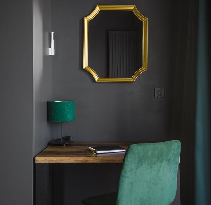 Kącik do pracy z zielonym aksamitnym fotelem, drewnianym biurkiem i złocistą ramą lustra na ciemnoszarej ścianie