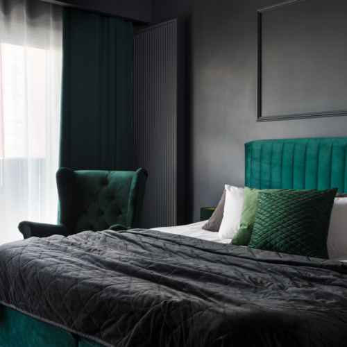 Elegancki pokój hotelowy z dużym łóżkiem z zielonym zagłówkiem i poduszkami, obok aksamitny fotel i zasłony przy oknie.