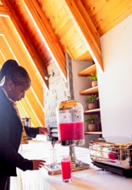 Guest in a cozy wooden lodge pouring bright pink juice from a beverage dispenser on a buffet table with plant-decor shelves nearby.