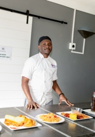 Chef in white uniform standing behind a counter with plated dishes and condiments in a modern hotel kitchen area