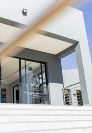 Modern hotel entrance with sleek glass doors, white steps, and a large artistic metal face sculpture under clear blue sky
