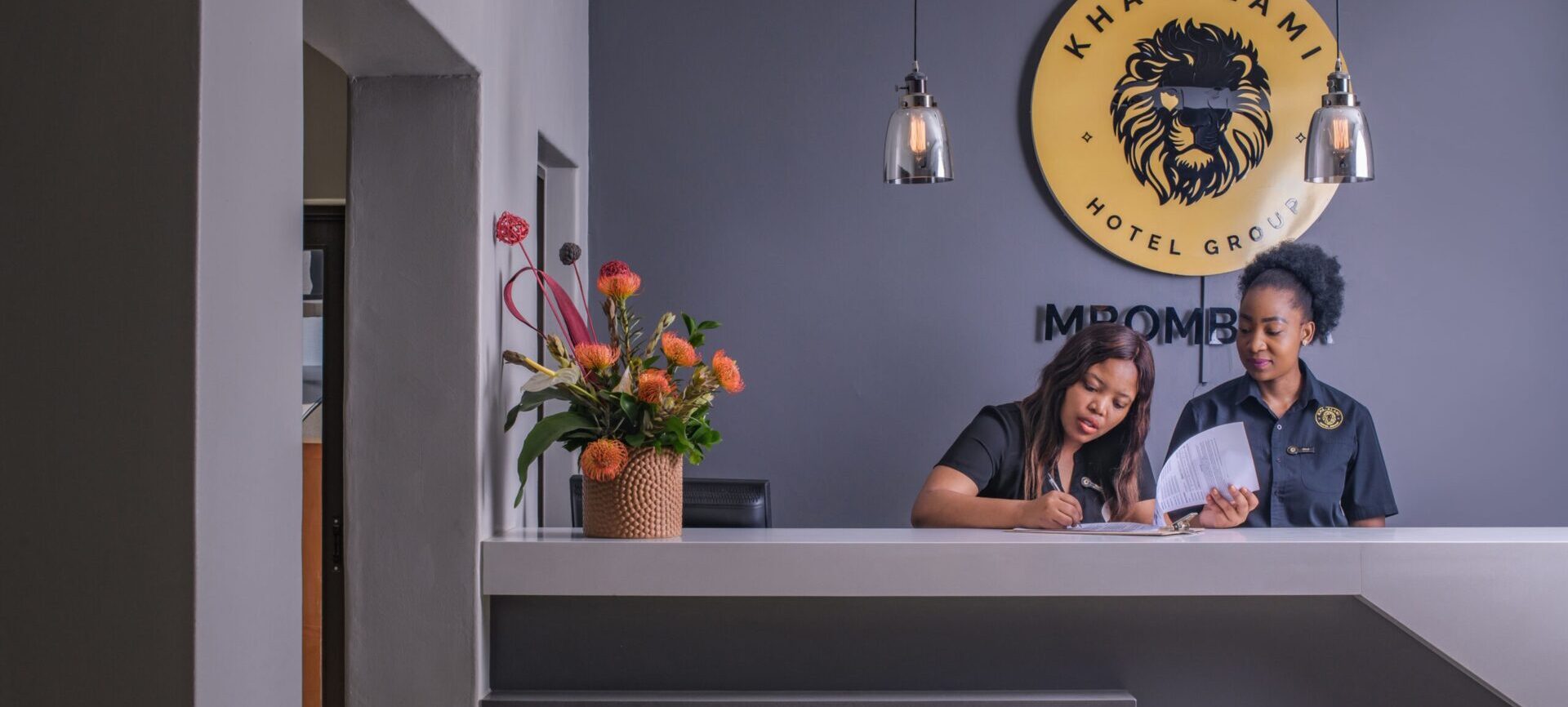 Hotel reception desk with two staff in black uniforms assisting with paperwork, modern decor, pendant lights, and vibrant floral arrangement
