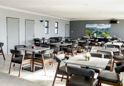 Bright, modern dining area with wooden tables and cushioned chairs, floral centerpieces, and a scenic landscape painting on the wall.