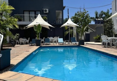 Outdoor swimming pool surrounded by lounge chairs with umbrellas, white patio furniture, and modern guest building under clear blue sky