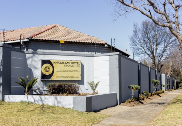 Khayalami Hotel Standerton exterior with a tiled roof, gated gray wall, landscaped plants, and a clear blue sky above