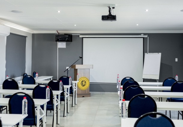 Modern hotel conference room with rows of white tables, navy chairs, bottled water, podium, projector, and pull-down screen