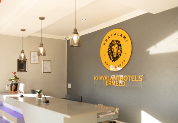 Modern hotel reception with sleek white desk, pendant lights, potted plants, and Khayalami Hotels Ermelo logo on gray wall
