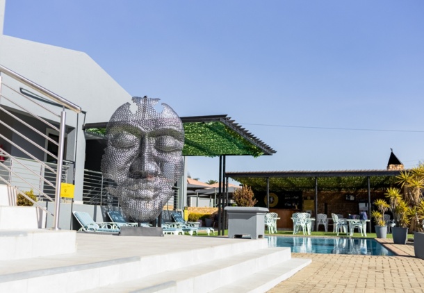 Outdoor hotel pool area with modern metal face sculpture, lounge chairs, shaded dining area, and tropical plants under clear blue sky