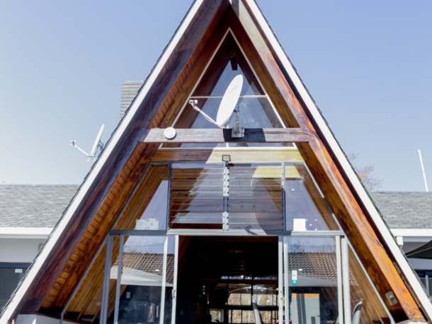 Modern A-frame hotel entrance with wooden beams, glass doors, and seating visible inside under clear blue sky