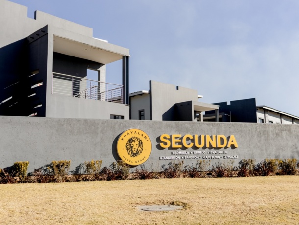 Modern hotel building with gray exterior and balconies behind a wall featuring Khayalami Hotel Group Secunda logo and signage