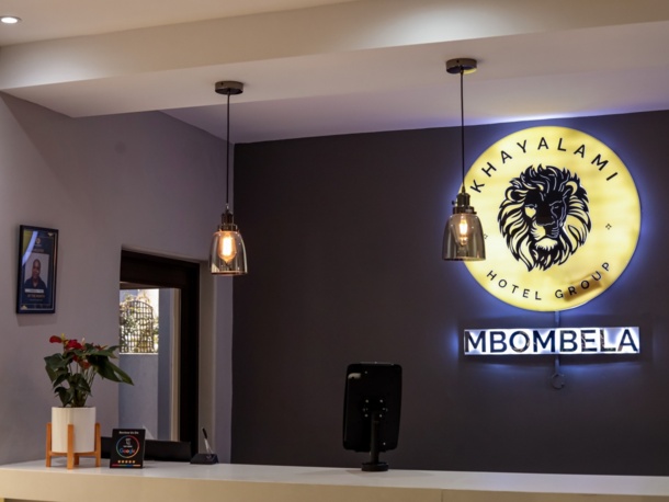Modern hotel reception with illuminated Khayalami Hotel Group Mbombela sign, pendant lights, and a small plant on white front desk