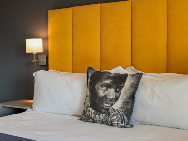 Modern hotel room with a yellow padded headboard, white linens, a decorative pillow with an artistic face, and safari-themed wall art above the bed