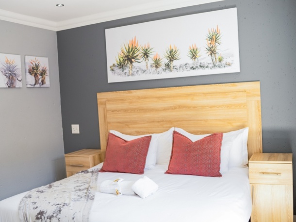 Cozy hotel room with a wooden headboard, white linens, two red accent pillows, matching nightstands, and botanical wall art