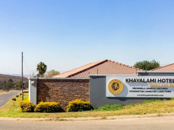 Khayalami Hotel Emalahleni entrance with brick and gray wall, terracotta roof tiles, and landscaped yellow bushes under clear blue sky