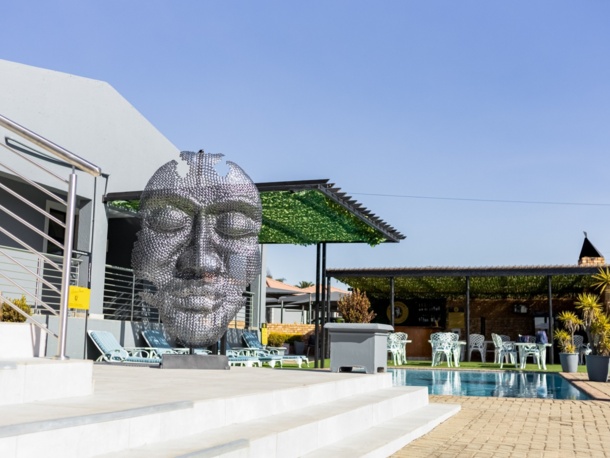 Outdoor hotel pool area with modern metal face sculpture, lounge chairs, shaded dining area, and tropical plants under clear blue sky