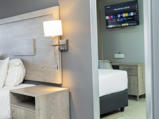 Modern hotel room corner with wooden nightstand, wall-mounted lamp, large mirror reflecting TV and part of the bed