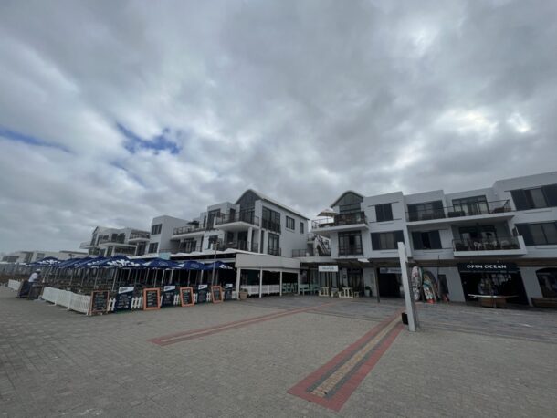 Coastal modern white hotel complex with multiple balconies, outdoor dining under blue umbrellas, and beachfront shops on a cloudy day