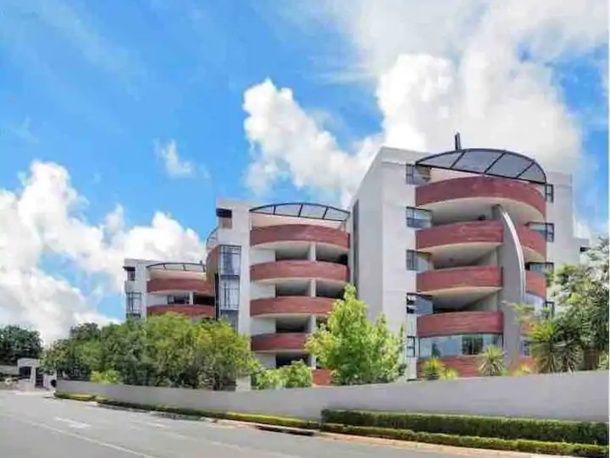 Modern hotel with curved balconies and glass railings, surrounded by lush greenery under a bright blue sky