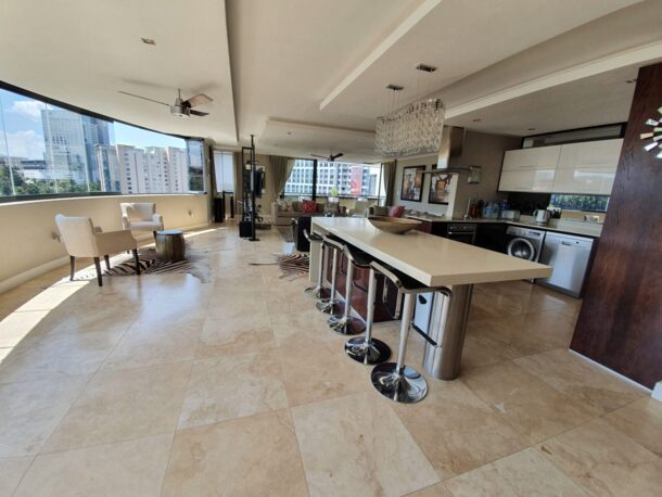 Spacious open-plan suite with modern kitchen island, bar stools, living area, large windows offering cityscape views, and natural light