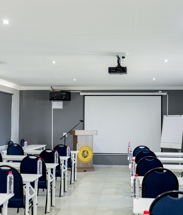 Modern hotel conference room with rows of white tables, navy chairs, bottled water, podium, projector, and pull-down screen