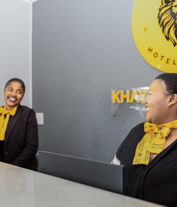 Two smiling hotel receptionists wearing black uniforms and yellow scarves at a modern front desk with a lion logo on the wall