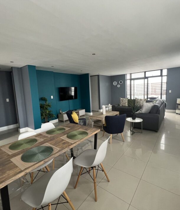 Spacious living and dining area with a wooden table, modern chairs, blue accent wall, flat-screen TV, and cozy seating near large windows
