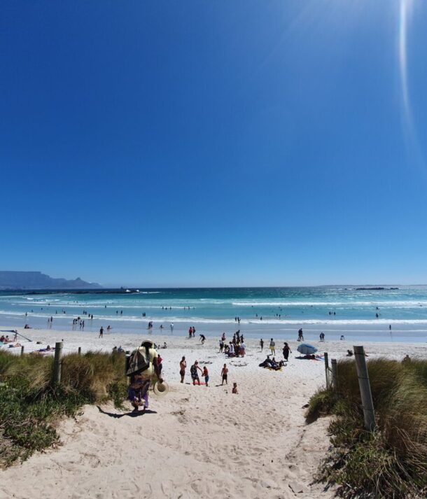 Sunny beach with white sand and people swimming, sunbathing, and playing, with a distant view of a flat-topped mountain under clear blue sky