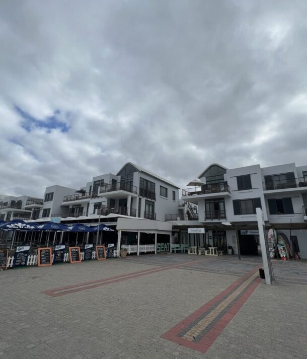 Coastal modern white hotel complex with multiple balconies, outdoor dining under blue umbrellas, and beachfront shops on a cloudy day