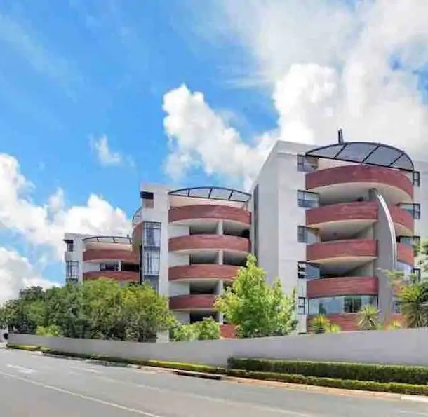Modern hotel with curved balconies and glass railings, surrounded by lush greenery under a bright blue sky