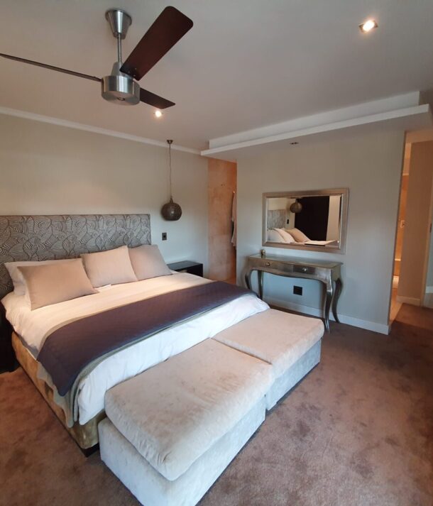 Cozy hotel bedroom with king bed, patterned headboard, hanging bedside lamps, bench seating, wall mirror, and neutral-toned decor