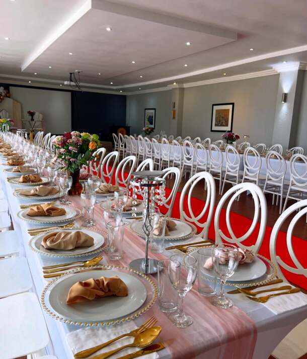 Elegant banquet room with long tables set for event, featuring gold utensils, beige napkins, crystal candle holders, and floral centerpieces