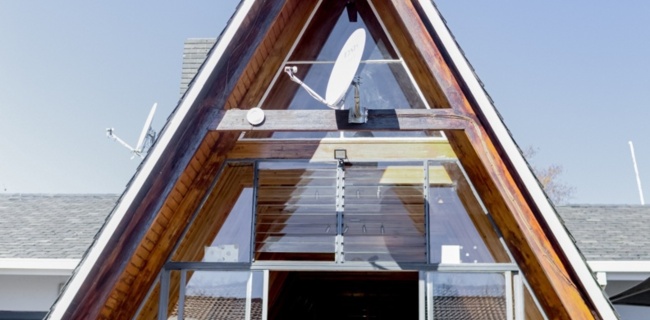 Modern A-frame hotel entrance with wooden beams, glass doors, and seating visible inside under clear blue sky