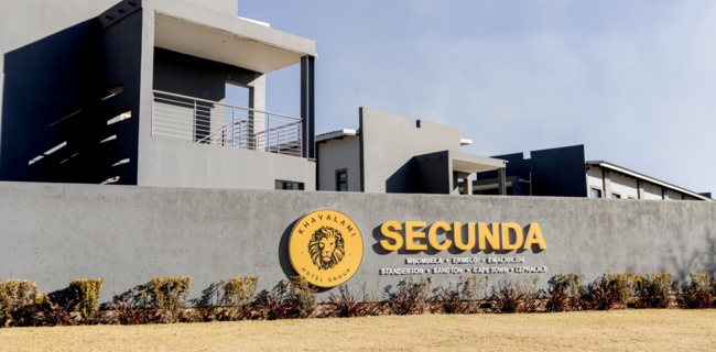 Modern hotel building with gray exterior and balconies behind a wall featuring Khayalami Hotel Group Secunda logo and signage