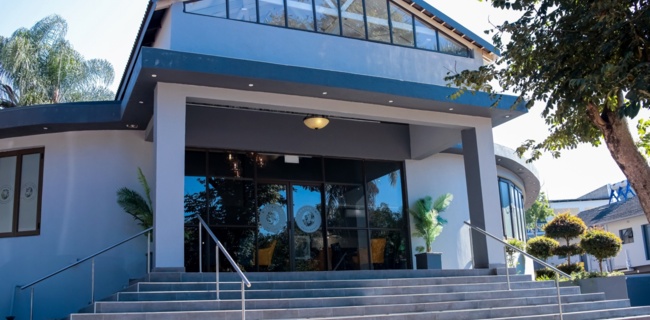 Modern hotel entrance with wide stairs, glass doors, potted plants, and a high triangular window under clear blue sky