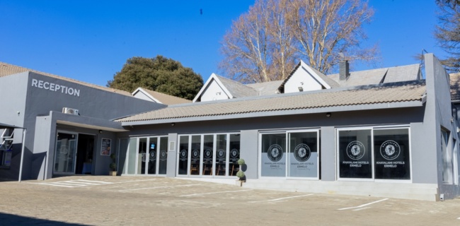Modern hotel reception building with large glass windows, clear signage, and paved parking area under a bright blue sky