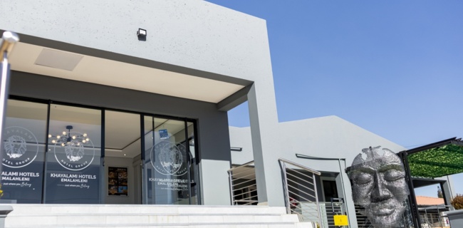 Modern hotel entrance with glass doors, wide white steps, and a large metal face sculpture under a clear blue sky