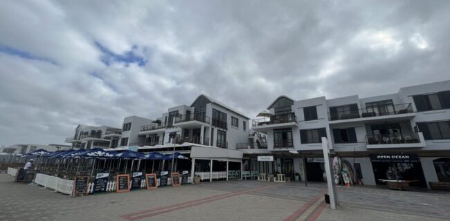 Coastal modern white hotel complex with multiple balconies, outdoor dining under blue umbrellas, and beachfront shops on a cloudy day