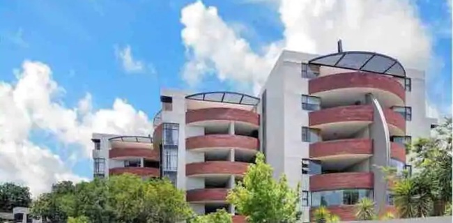 Modern hotel with curved balconies and glass railings, surrounded by lush greenery under a bright blue sky