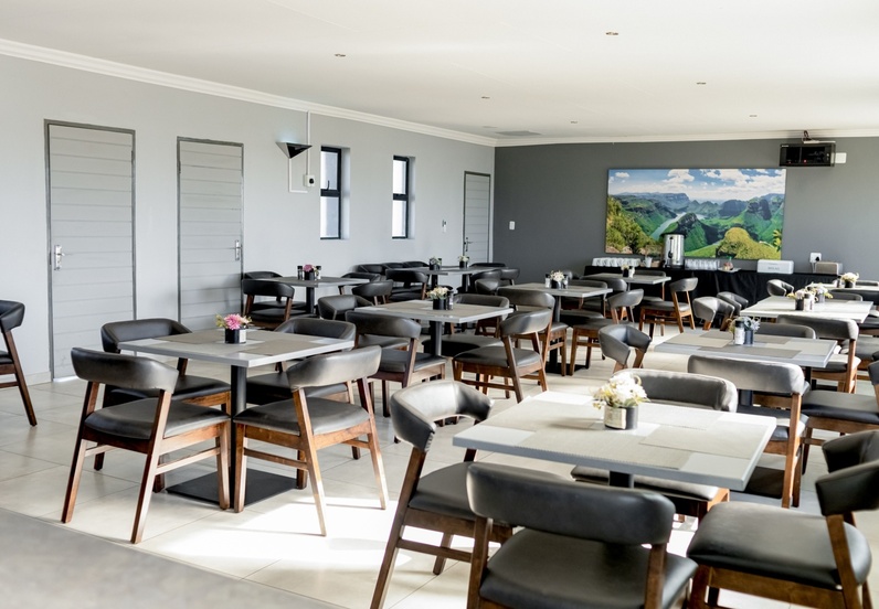 Bright, modern dining area with wooden tables and cushioned chairs, floral centerpieces, and a scenic landscape painting on the wall.